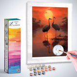 Paint by Numbers kit of two cranes at sunset with a warm red and golden sky reflecting on the water, evoking harmony and peace.