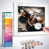 Paint by Numbers kit featuring a violin on aged music paper, bathed in morning light, with numbered paint pots and a hand holding a brush.